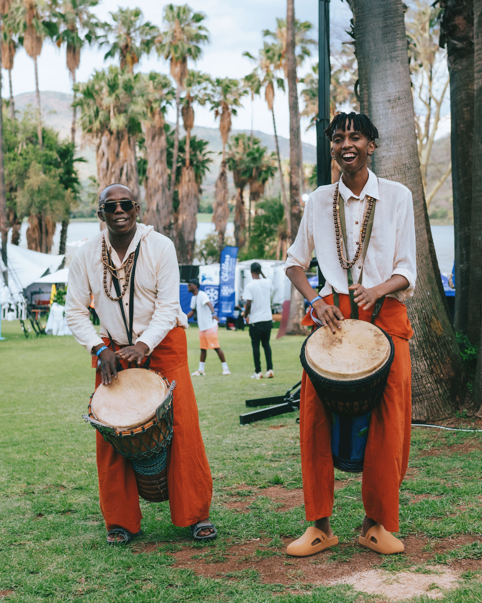 Two performers in orange pants playing djembe drums outdoors at the day event