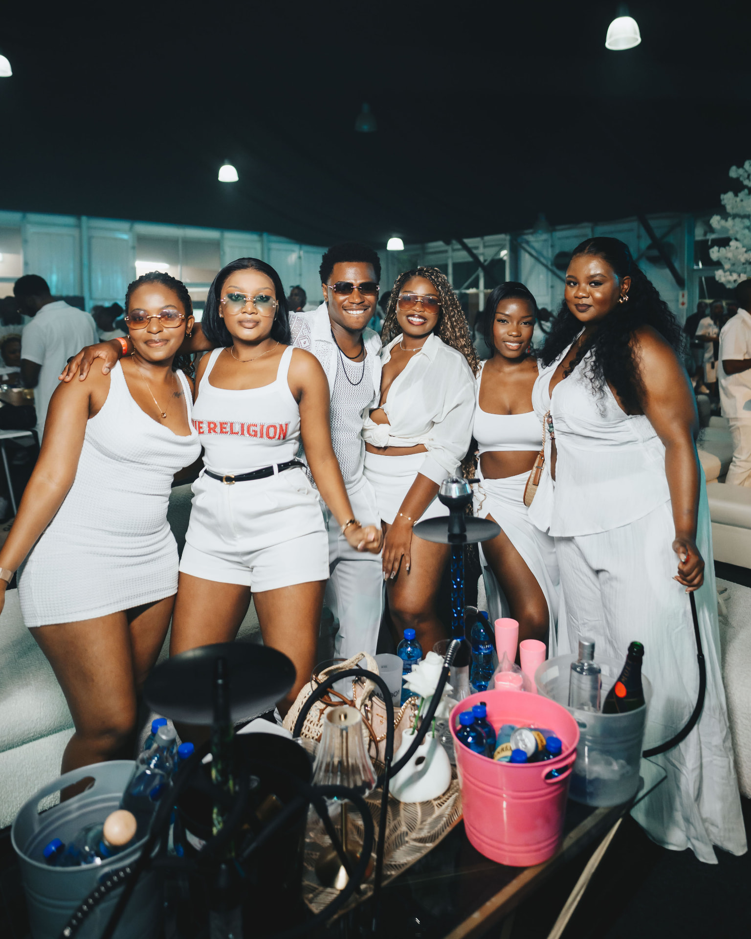 Six guests in all-white outfits posing around a table with bottle service